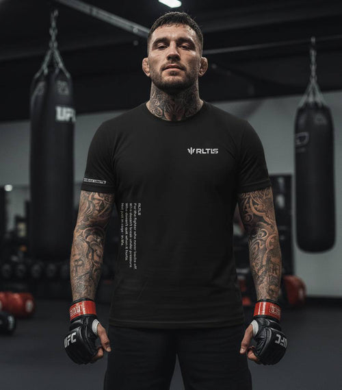 Man in a black Altis t-shirt with tattoos standing in a boxing ring.